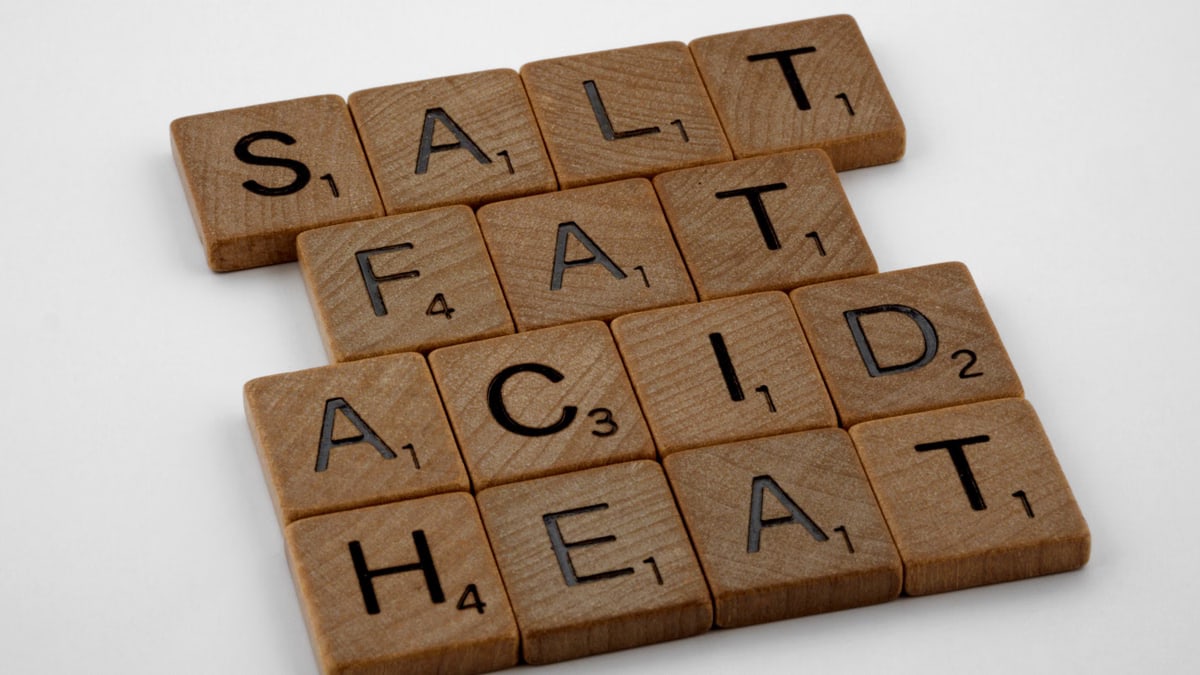 Arrange of wooden letter tiles spelling 'Salt Fat Acid Heat' on a white background.